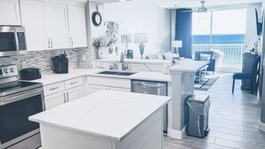 Eine moderne Küche mit weißen Schränken, einer Mikrowelle, einem Toaster und einem Spülbecken. Es gibt einen Essbereich mit Tisch und Stühlen, und durch ein großes Fenster blickt man auf den Ozean.