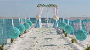 Hochzeitszeremonie am Strand mit einem weißen Bogen, blauen und weißen Blumenschmuck und einem sandigen Weg zum Altar.