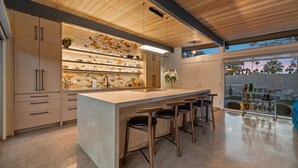 Una cocina moderna con una isla central, taburetes de bar, armarios de madera y vistas al exterior.