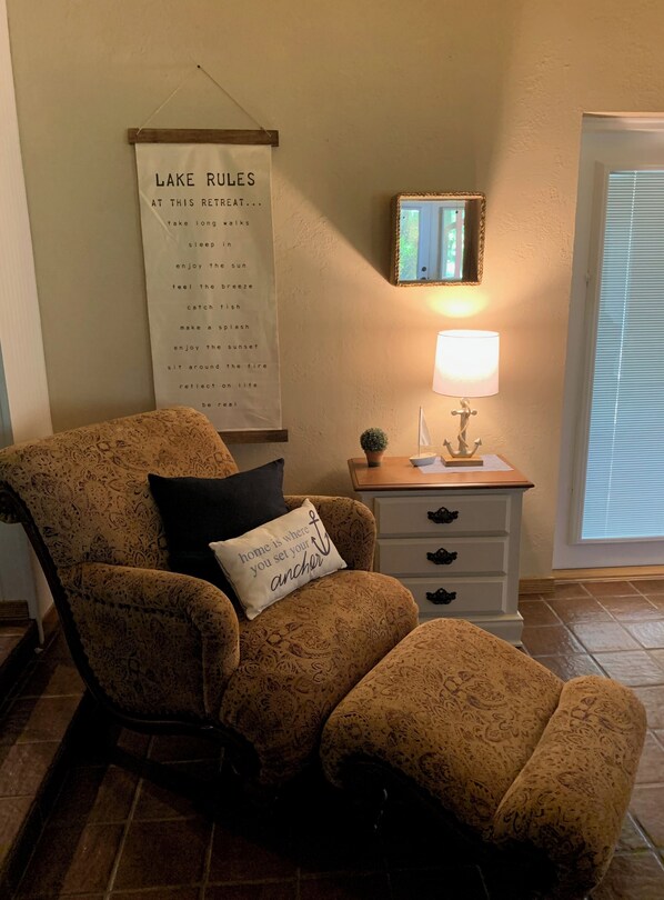 Un salon avec un fauteuil à motifs, une petite table avec une lampe, un tableau encadré et une affiche accrochée au mur.