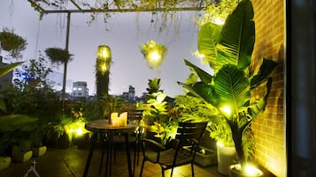 Une terrasse extérieure avec une table et des chaises, entourée de plantes en pot et de plantes suspendues, avec une vue sur la ville en arrière-plan.