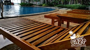 Un área junto a la piscina con tumbonas de madera, palmeras y un edificio al fondo.