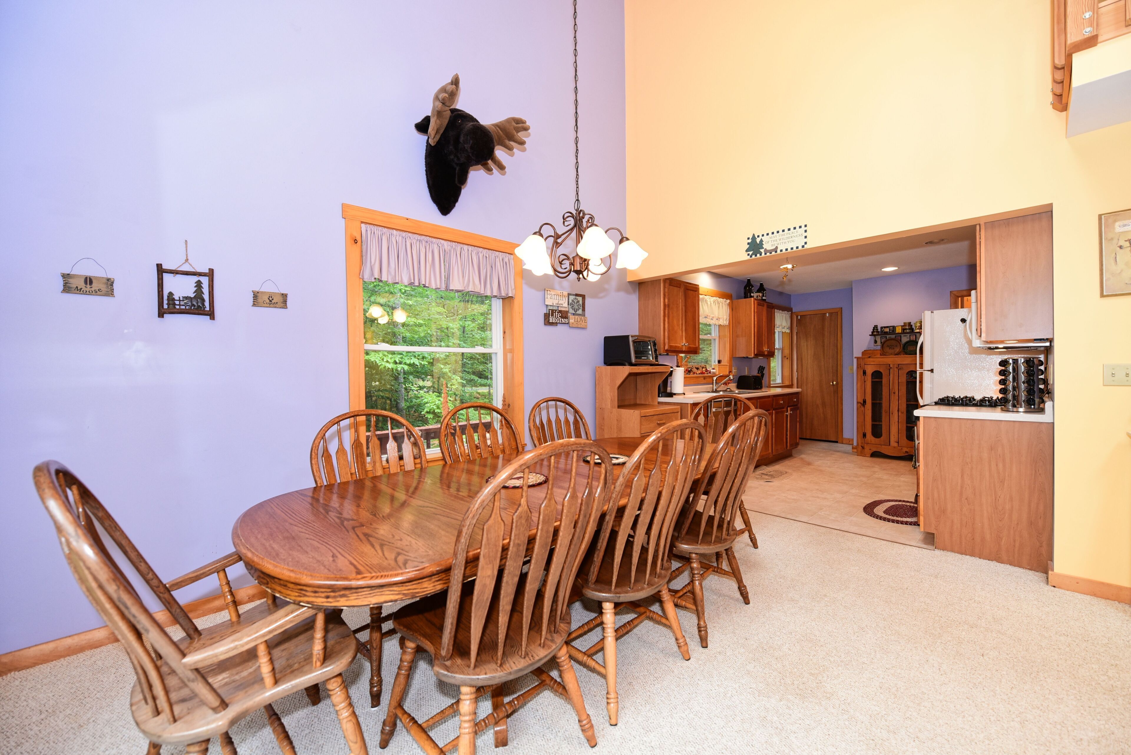 A dining room with a wooden table and chairs, a mounted animal head on the wall, and a view of the kitchen and living area.