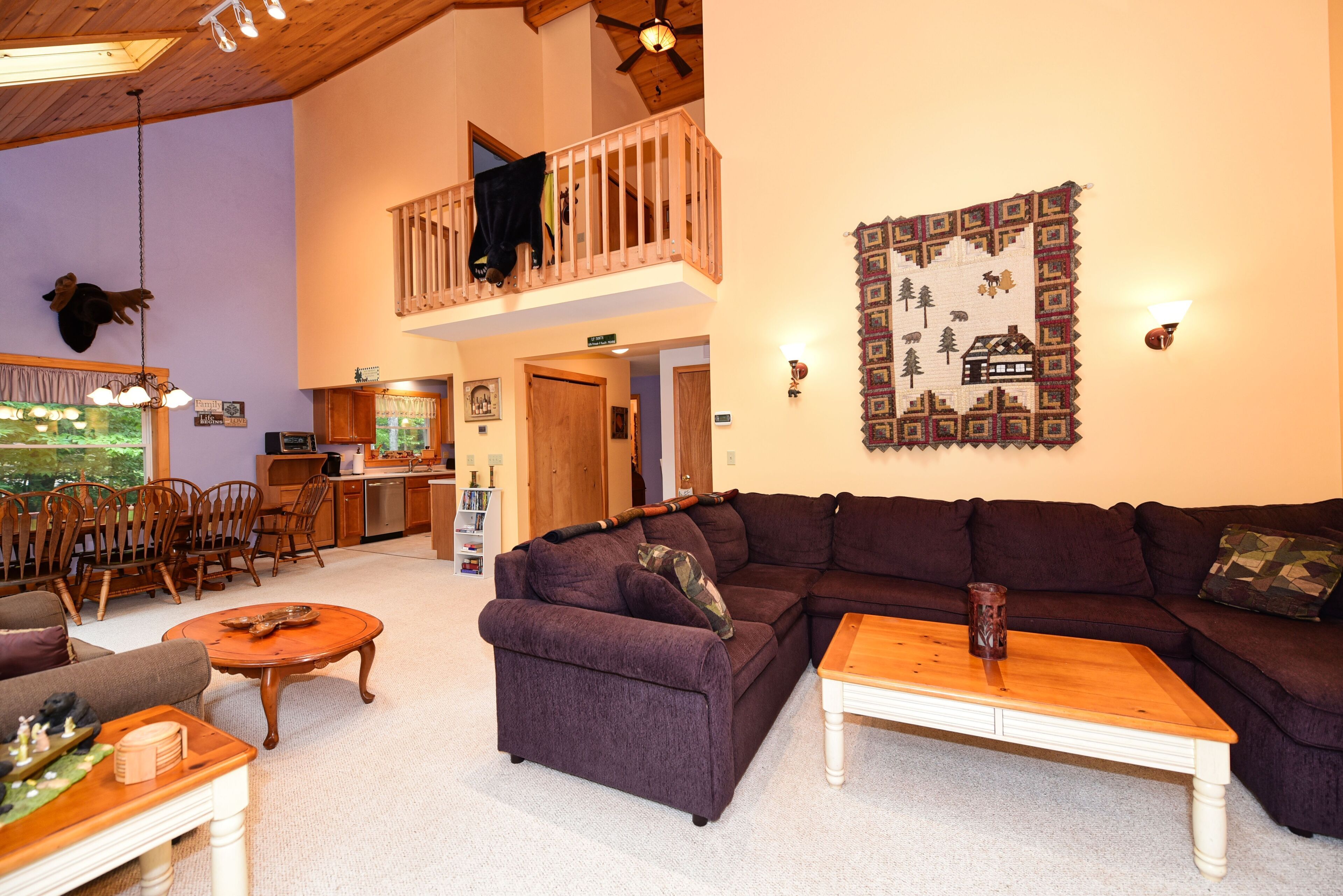 A spacious living area with a large sectional sofa, a wooden coffee table, and a dining area with wooden chairs and a table. A balcony with a railing is visible, and a decorative wall hanging is present.