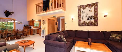 A spacious living area with a large sectional sofa, a wooden coffee table, and a dining area with wooden chairs and a table. A balcony with a railing is visible, and a decorative wall hanging is present.