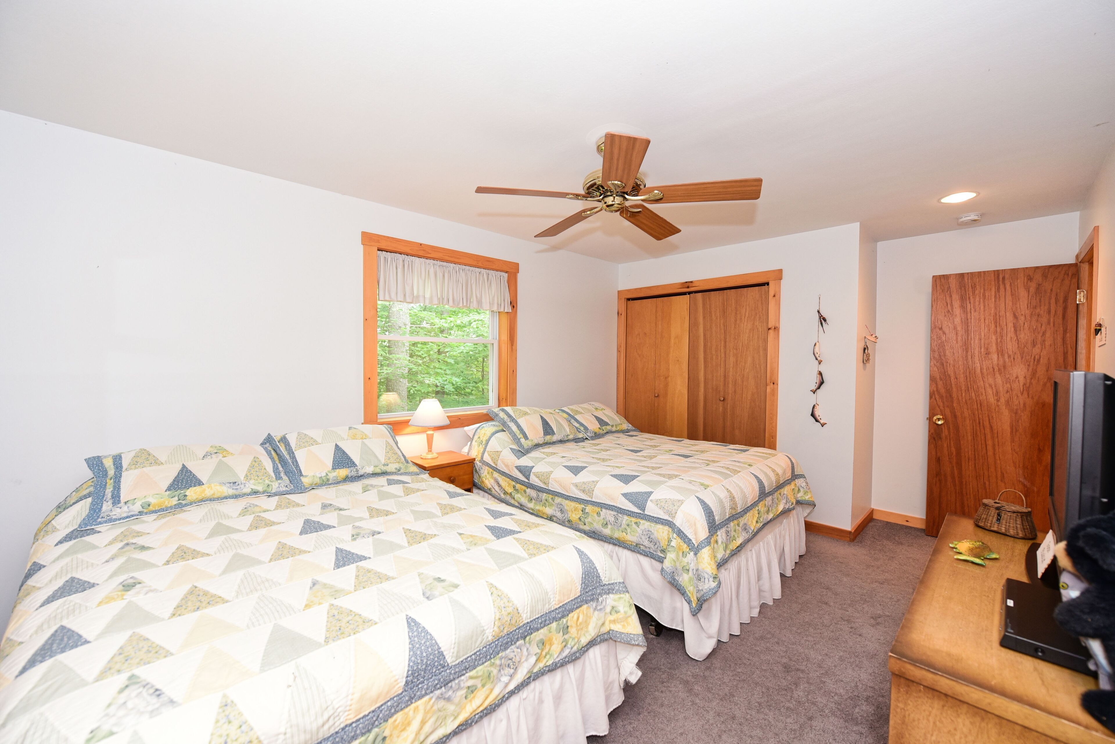 A bedroom with two beds, a ceiling fan, a window with a view of greenery, and a wooden door.