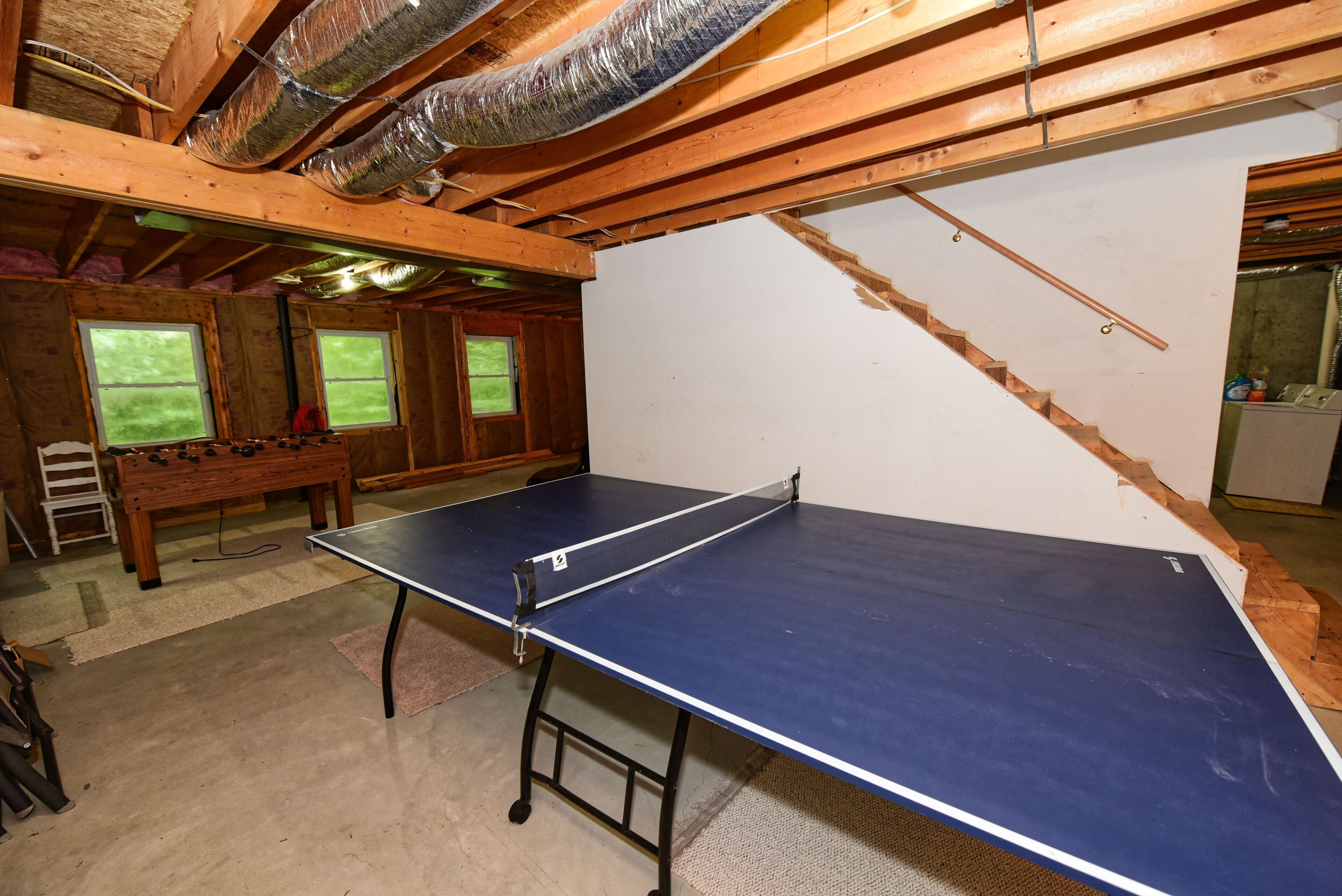 A room with a ping pong table, a foosball table, and a window with a view of trees.