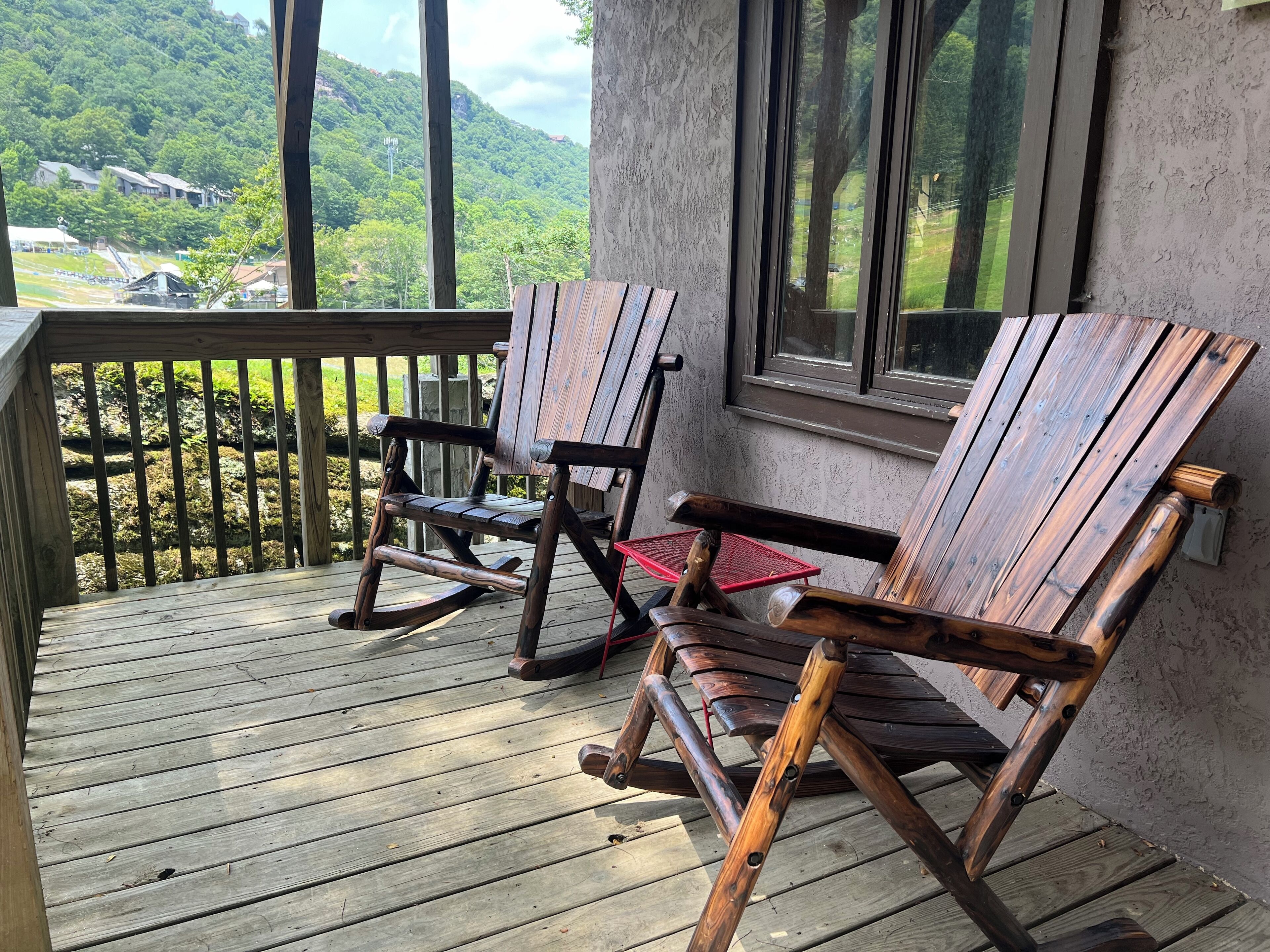 Eine Holzterrasse mit zwei Schaukelstühlen und einem roten Tisch, mit Blick auf eine grüne Landschaft mit Hügeln und Gebäuden.