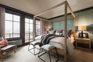 A four-poster bed with a fur throw, two bedside lamps, a wooden nightstand, and a view of the outdoors through large windows.