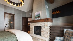 A bedroom with a stone fireplace, a bed with a green blanket, a plaid sofa, and a mounted TV. - Silver Star 40 | Theater Room | Hot Tub (Park City)