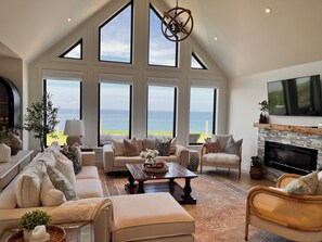 Un salon spacieux avec un haut plafond, de grandes fenêtres donnant sur la mer, une cheminée et des sièges confortables.