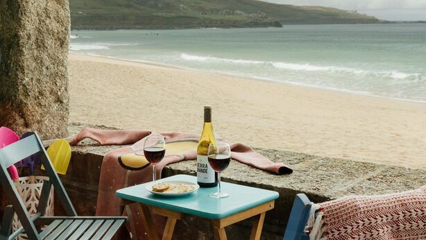 A beachside table with a bottle of wine, glasses, and a plate of food, overlooking the ocean. - 6 Barnaloft (St Ives)
