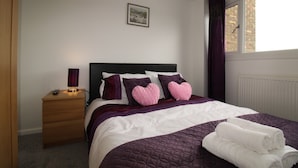 A neatly made bed with a purple quilt, two heart-shaped pillows, and rolled white towels.