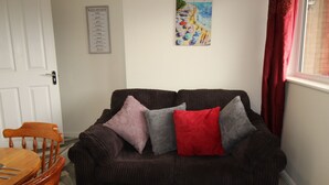 A dark brown sofa with four cushions in shades of purple, gray, and red, positioned against a white wall with a poster titled "La Plage".