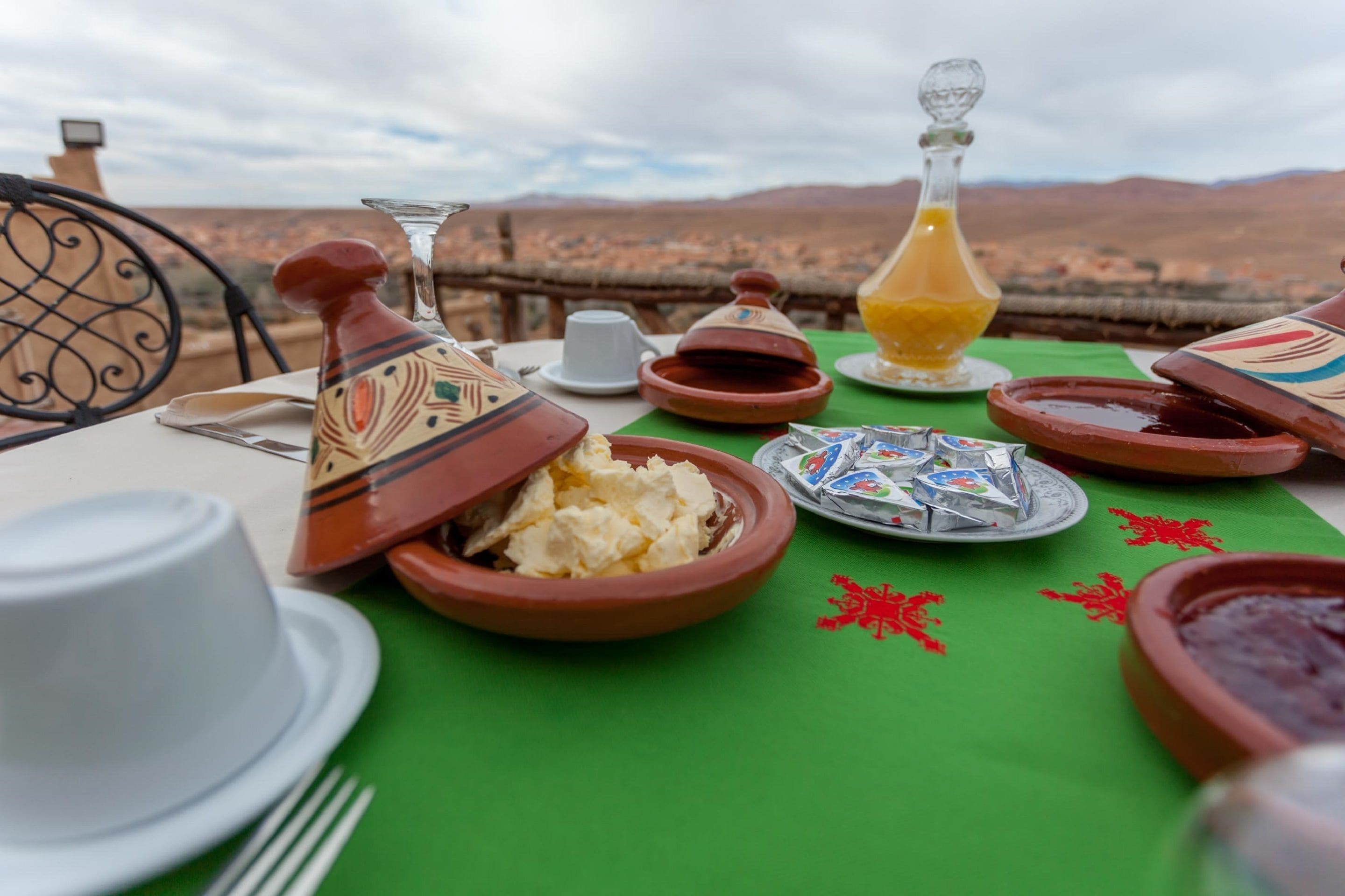A table set with traditional Moroccan dishes, including a tagine, a jug of orange juice, and a plate of pastries, overlooking a mountainous landscape.