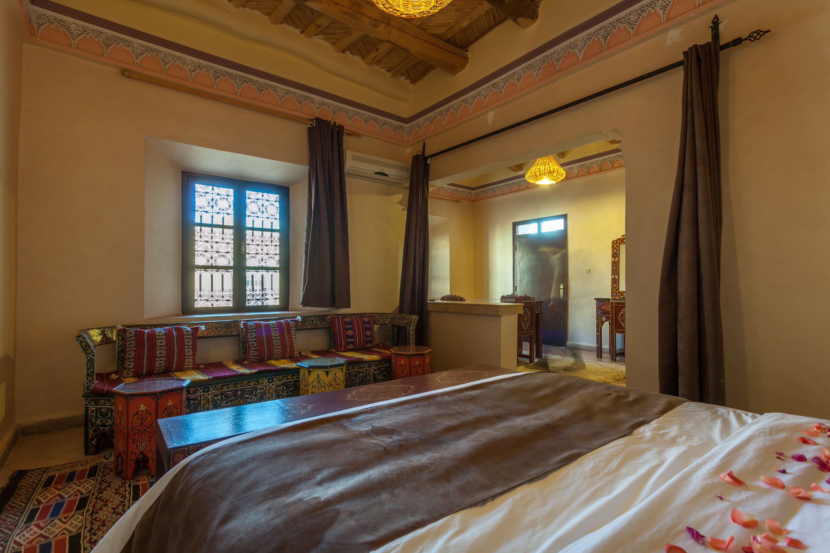 Una habitación tradicional con una cama grande, un banco con cojines coloridos y una ventana con rejas decorativas de hierro.