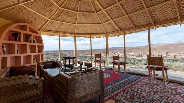 Ein Zimmer mit einem Strohdach, Holzmöbeln und Blick auf eine Wüstenlandschaft.