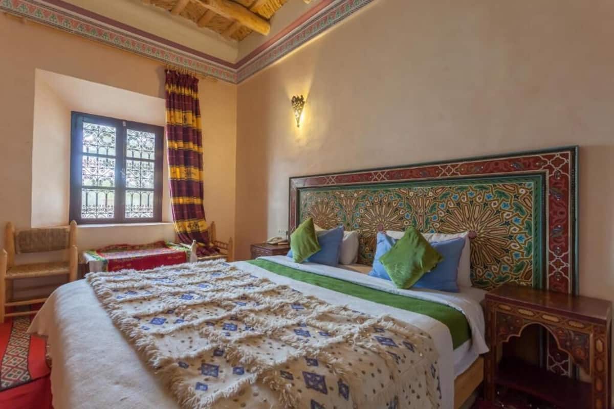 Une chambre à coucher avec un grand lit, une fenêtre avec des rideaux, une chaise et une tête de lit décorative.