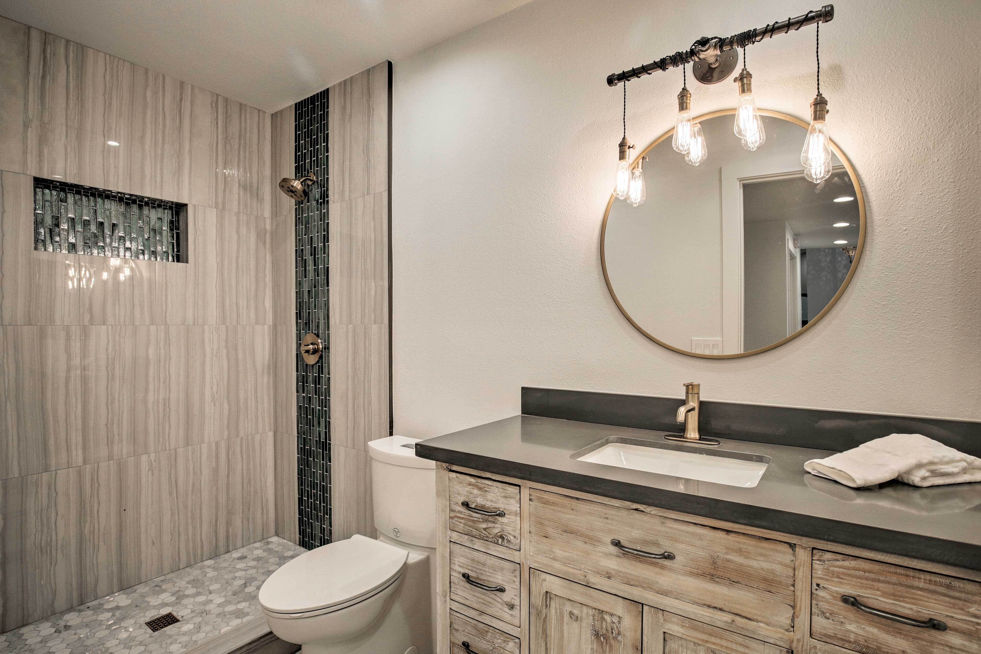 A modern bathroom with a marble shower wall, a toilet, a sink with a wooden vanity, and a mirror with hanging lights.