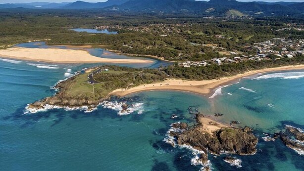 Vista aérea de una zona costera con playa, afloramientos rocosos y un pueblo al fondo.
