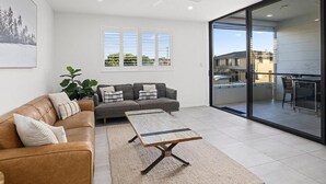 Un salón moderno con un sofá de cuero marrón, un sofá gris y una mesa de centro. Se aprecia una ventana grande con persianas, una planta y vistas a un balcón.