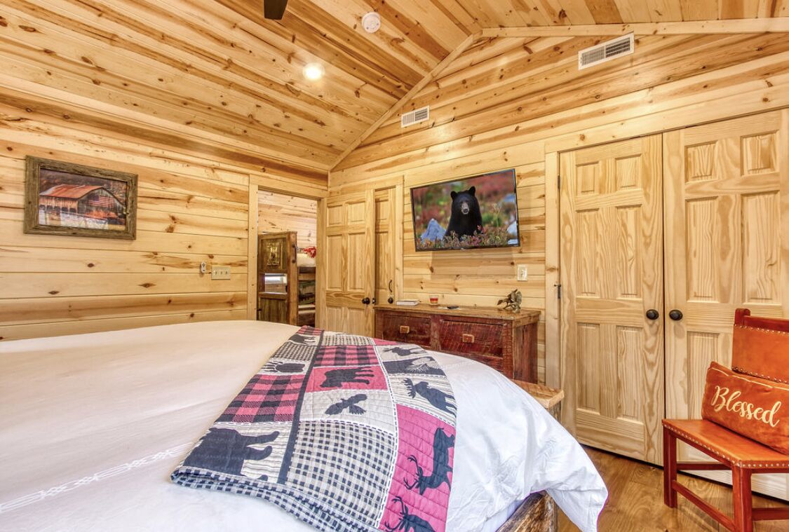 Une chambre dans un chalet en bois, avec un lit, une télévision et un fauteuil agrémenté d'un coussin orné de la mention « Béni ».