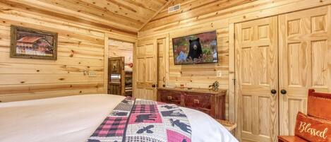 Une chambre dans un chalet en bois, avec un lit, une télévision et un fauteuil agrémenté d'un coussin orné de la mention « Béni ».