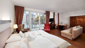 A hotel room with a large bed, a sofa, a desk with a chair, a TV, and a view of buildings through large windows.