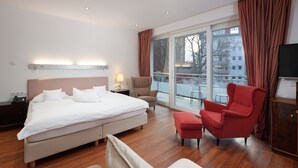 A hotel room with a large bed, a red armchair, a small ottoman, a TV, and a balcony with a view of buildings.