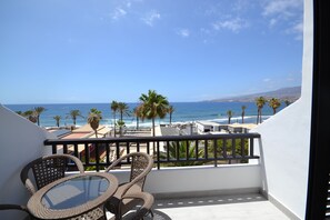 A balcony with wicker chairs and a glass table overlooking a beach with palm trees and the ocean. - Hermoso tramo de costa (Playa de la Américas)