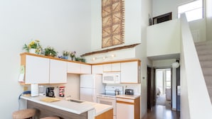 A modern kitchen with white cabinets, a microwave, a stove, and a sink. There are two stools and a small island with a cutting board.