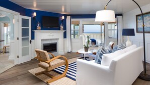 Sala de estar com sofá branco, cadeira de balanço de madeira, lareira e uma janela grande com vista para o oceano.