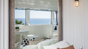 Quarto com uma grande janela com vista para o mar, cama com várias almofadas e uma pequena mesa com uma planta.