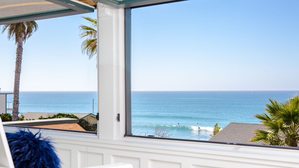 Quarto com uma grande janela que dá para uma praia com palmeiras e o oceano.