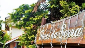 Ein hölzernes Schild mit dem Schriftzug 'Houses da Serra' in einem Garten mit violetten Blumen und Grünpflanzen.