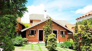 Ein zweistöckiges Haus mit Ziegeldach, Schornstein und einer Veranda. Auf der Veranda stehen Kübelpflanzen und vor dem Haus ein gepflegter Rasen.