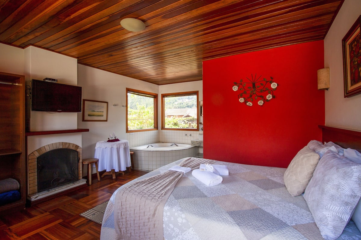 Ein Schlafzimmer mit Kamin, einem großen Bett, einer roten Akzentwand und Blick ins Freie durch ein Fenster.