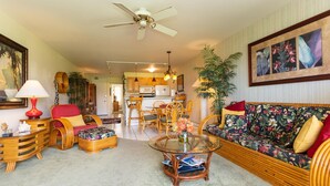 A living room with a glass coffee table, wicker furniture, a sofa with floral cushions, and a kitchen in the background. - Hale Kamaole 220 Second floor condo, easy access to best beach on Maui! (Kihei)