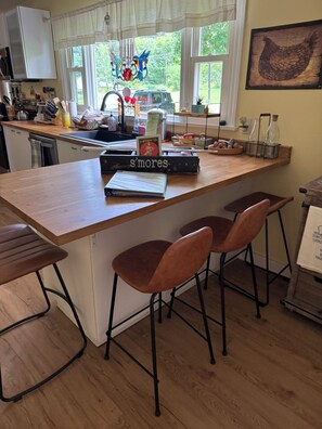 A kitchen with a wooden countertop, a sink, and a window with a view of greenery. There are two stools and a book titled 's'mores' on the counter. - Lovely ranch home getaway in The Berkshires- just 6 miles to Tanglewood (Richmond)
