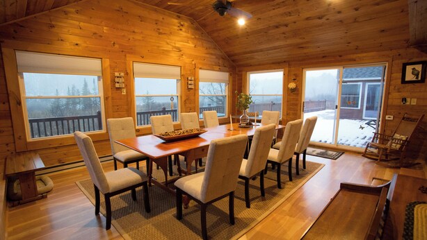 Interior de una cabaña de madera con una mesa de comedor, sillas y ventanas amplias.