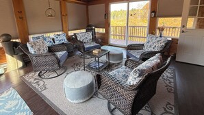 Un interior de madera acogedor con muebles de mimbre, una mesa central y vistas al exterior a través de amplios ventanales correderas.