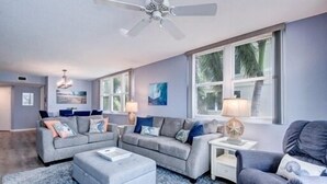 Ein modernes Wohnzimmer mit einer grauen Sofagarnitur, einem blauen Sessel und einem weißen Hocker. Es gibt einen Deckenventilator, ein Fenster mit Blick auf Palmen und eine Lampe auf einem Beistelltisch.