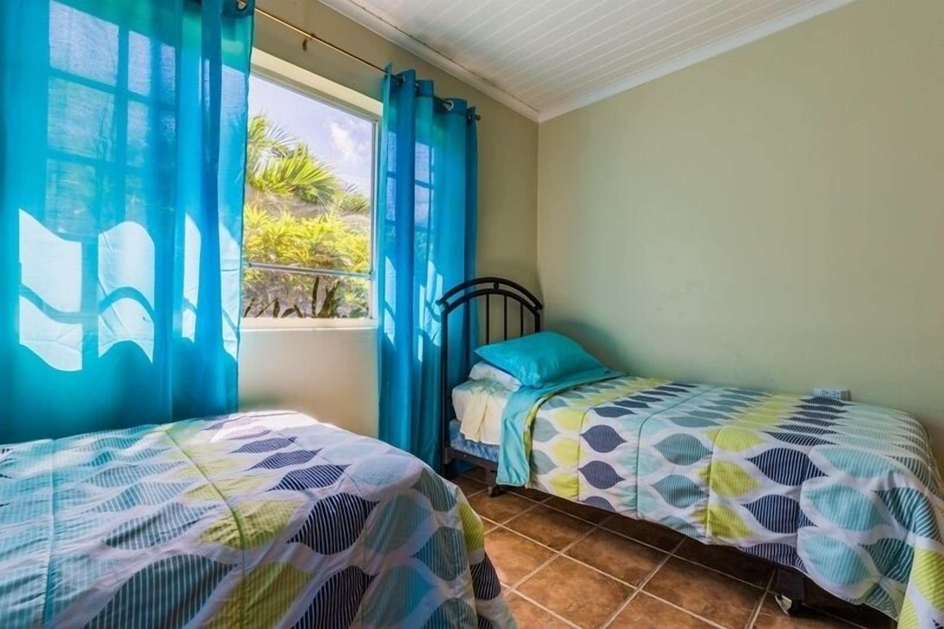 Ein Zimmer mit zwei Betten, jeweils mit gemusterten Bettbezügen, einem Fenster mit blauen Vorhängen und Blick auf Grünfläche.