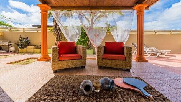 A covered outdoor seating area with wicker chairs and a table, a pool, and a view of the sky. - Palm Beach home with private pool (Noord)