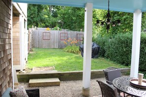 Eine überdachte Terrasse mit Tisch und Stühlen, eine Holzterrasse und ein eingezäunter Garten mit Rasen und Blumenbeeten.