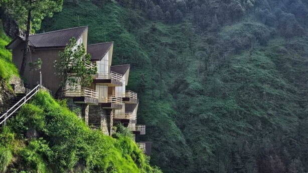 A modern building with multiple balconies, situated on a hillside with lush greenery and a forested background.