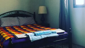 Ein Schlafzimmer mit Bett, Nachttisch, Lampe, einem Fenster mit Vorhängen und einem Bild an der Wand.