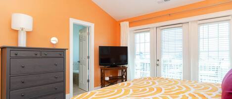 A bedroom with an orange wall, a bed with a yellow and white patterned bedspread, a dark wooden dresser with drawers, a flat-screen TV on a stand, and a door leading to another room.