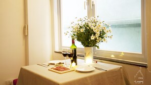 Une table dressée avec une bouteille de vin, des verres, une assiette de nourriture et un vase de fleurs, placée près d’une fenêtre donnant sur la mer.