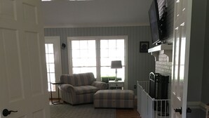 Ein Wohnzimmer mit einer Couch, einem Hocker und einem gemusterten Teppich. Eine Tür führt in einen anderen Raum, ein Fenster mit Jalousien und ein Fernseher auf einem Schrank.